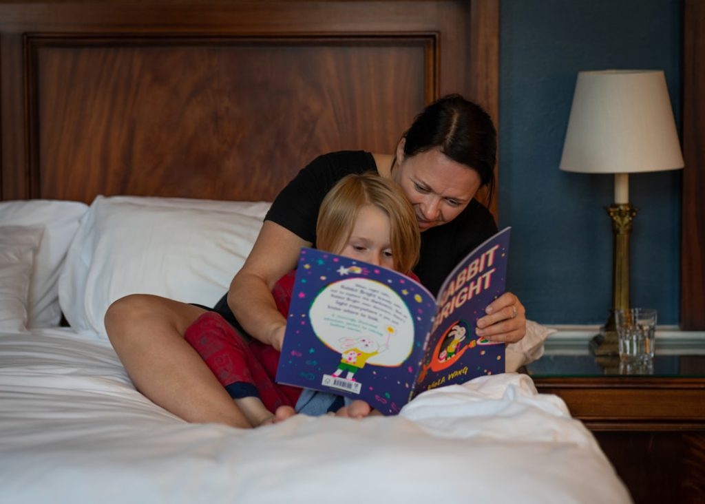 Mother reading a bedtime storybook to her young child on a cozy bed with a lamp in the background