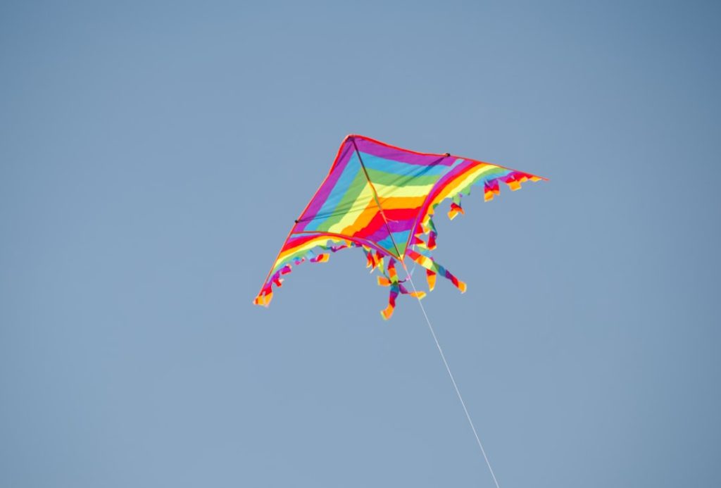 A colorful rainbow diamond kite with a long tail soaring high against a clear blue sky