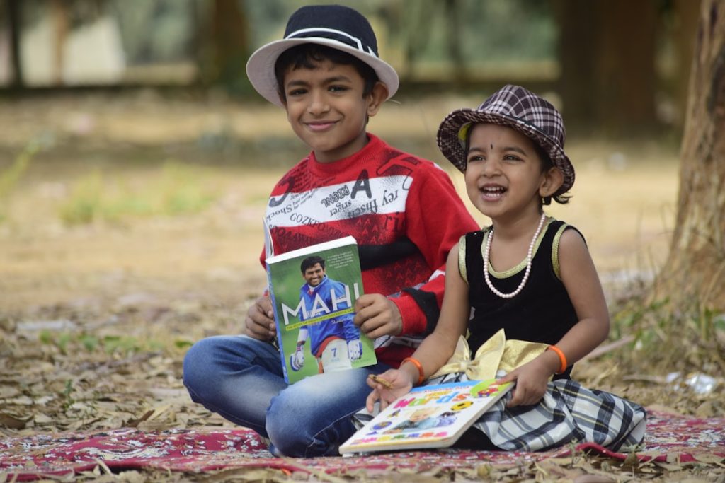 Two South Asian children sitting outdoors holding colourful storybooks and smiling