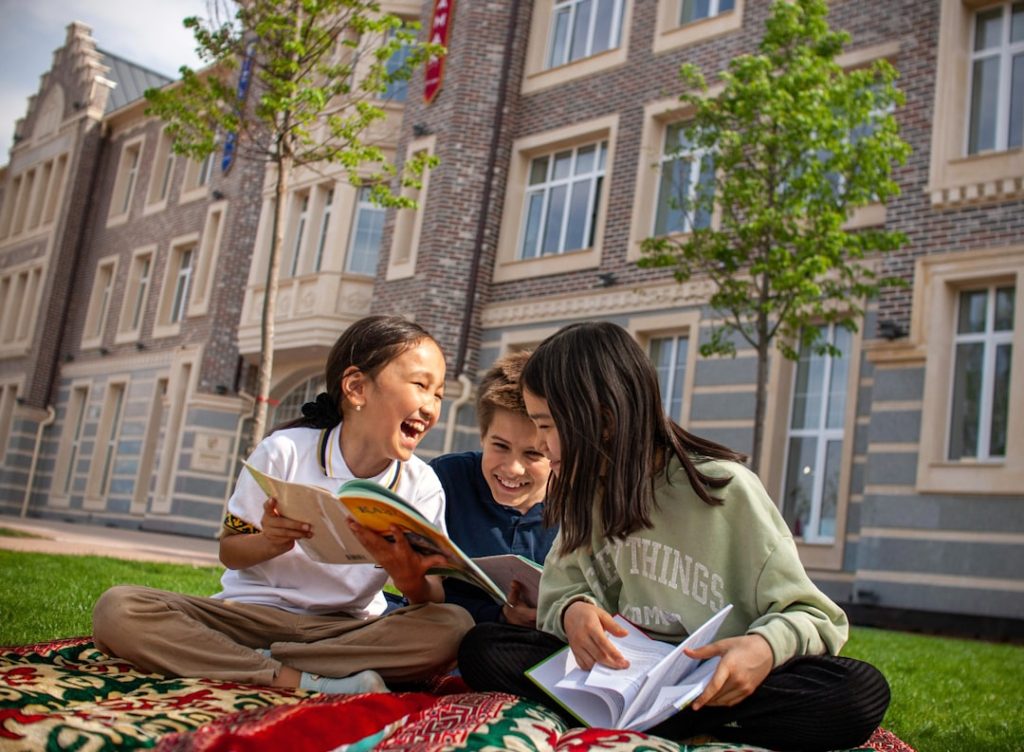 Three children sitting on grass outside a school building, laughing together while reading books