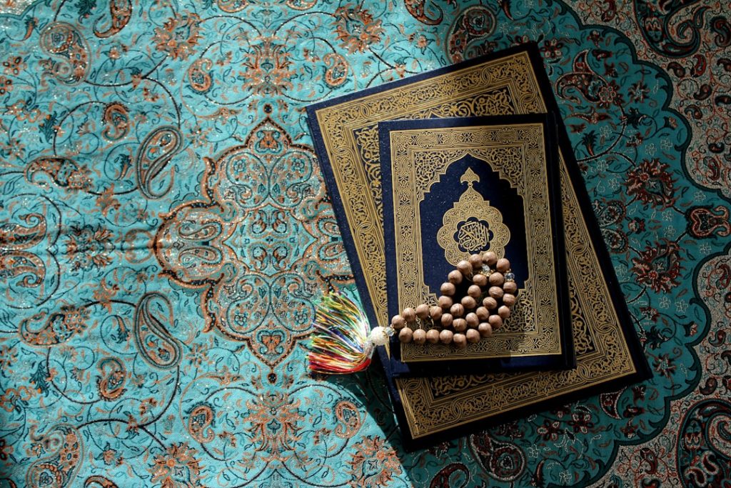 Holy Quran with wooden tasbih prayer beads resting on top, placed on a decorative teal patterned cloth